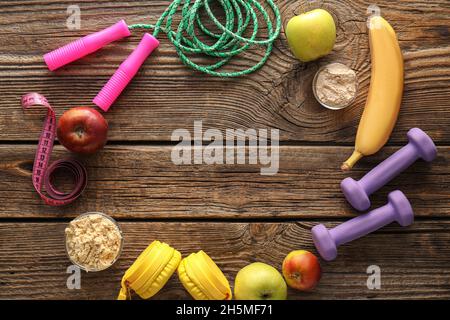 Telaio in ciotole con polvere proteica, frutta, manubri, corda da salto, nastro di misurazione e cuffie su sfondo di legno Foto Stock