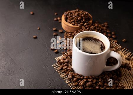 Tazza bianca con caffè caldo e chicchi di caffè tostati sparsi su un rustico tavolo nero. Spazio di copia per il testo Foto Stock