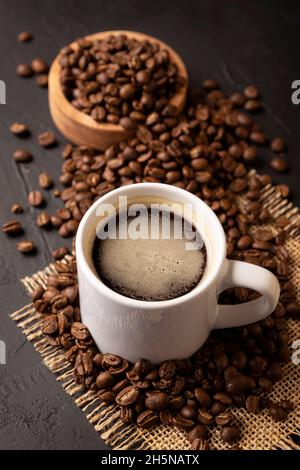 Chicchi di caffè sparsi su un rustico tavolo nero e una tazza bianca con caffè caldo. Immagine da vicino Foto Stock