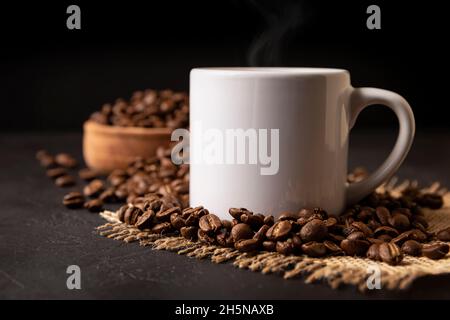 Tazza bianca con caffè caldo e chicchi di caffè tostati sparsi su un rustico tavolo nero. Immagine da vicino. Tazza vuota per spazio di copia Foto Stock