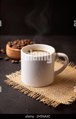 Tazza bianca con caffè caldo e chicchi di caffè tostati sparsi su un rustico tavolo nero. Immagine da vicino. Tazza vuota per spazio di copia Foto Stock