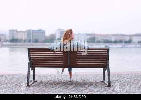 Giovane donna rossa seduta su panca in riva al fiume vista posteriore Foto Stock