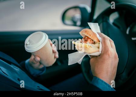 Uomo d'affari che fa colazione all'interno dell'auto. L'autista sta pranzando. Fast food, cibo spazzatura in mano di un giovane uomo d'affari in un vestito. Foto Stock