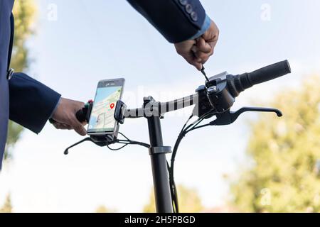 Primo piano di un uomo che utilizza uno smartphone per noleggiare un veicolo e-kick. Ragazzo millenario con telefono cellulare che utilizza il trasporto ecologico in centro. Stile di vita urbano moderno i Foto Stock