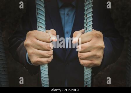 Giovane uomo d'affari corrotto in prigione, dietro i bar della prigione. Le mani degli uomini tengono una griglia rinforzata al buio. Il concetto di crimini finanziari Foto Stock