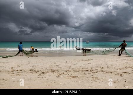 I pescatori tirano le reti dal mare su una spiaggia nei tropici con il cielo drammatico su Mahe Island, Seychelles Foto Stock