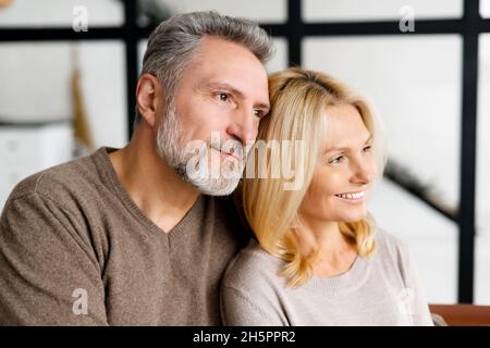 I coniugi di mezza età siedono sul divano a casa e guardano da parte in un senso. Primo piano ritratto di armoniosa coppia felice, uomo bello dai capelli grigi e affascinante donna bionda Foto Stock
