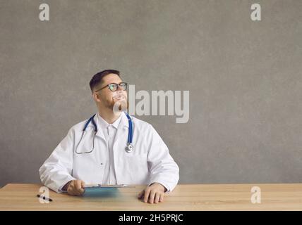 Medico contemplativo soddisfatto seduto alla scrivania tenendo appunti studio scatto Foto Stock
