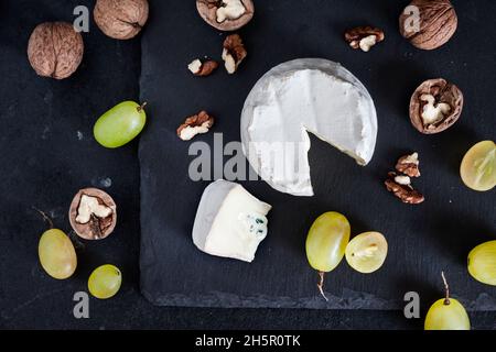 Tipo di formaggio Brie. Formaggio morbido con uva e noci su sfondo nero. Disposizione piatta, vista dall'alto Foto Stock