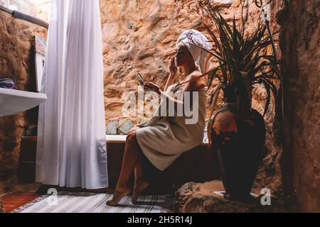 Felice elegante donna adulta hanno cura e fare il trattamento di bellezza della pelle a casa in bagno di design rurale. Donne e attività di cura della pelle a casa Foto Stock