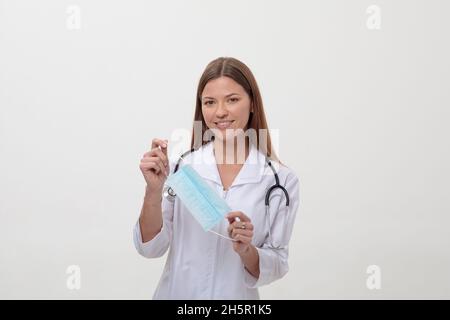 Una giovane dottoressa in uniforme tiene una maschera medica su sfondo bianco. L'infermiere mostra il pollice ok. Foto Stock