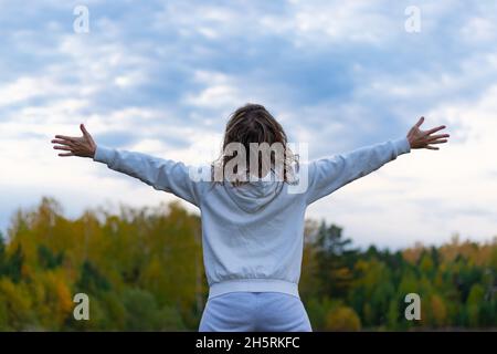 Una donna in un maglione bianco con un cappuccio si erge in un campo sullo sfondo di una foresta e un cielo blu con le braccia estese su un caldo a Foto Stock