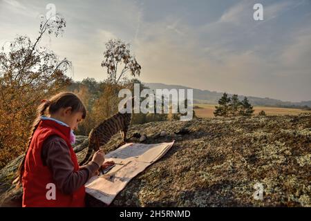 BLATNO, REPUBBLICA CECA - CIRCA OTTOBRE 2021: Una bambina si occupa di scrivere calligrafia all'esterno sulle rocce e il suo gatto osserva circondare. Foto Stock