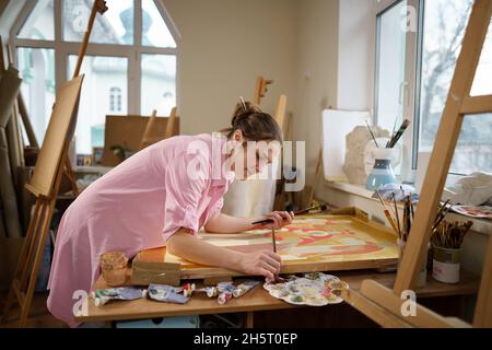 Cute donna dipinge su tela in un laboratorio d'arte. Artista che crea l'immagine. Scuola d'arte o studio. Lavorare con vernici, spazzole e cavalletto. Hobby e tempo libero Foto Stock