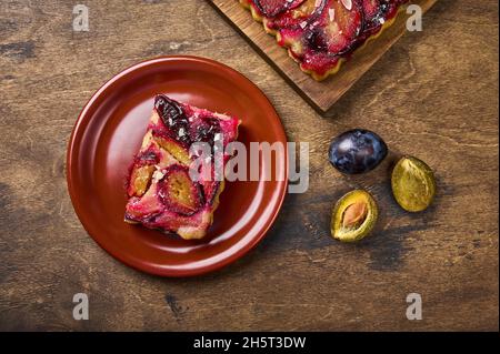 Pezzo fatto in casa di crostata di susina su piastra di ceramica, accanto a e le metà di susina e torta su tavola di legno. Vista dall'alto in stile rustico, spazio copia Foto Stock