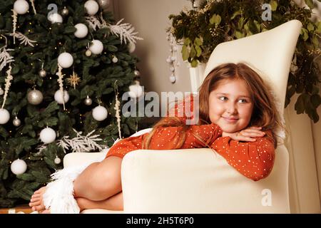 Giovane felice divertente sorridente sognando ragazza dai capelli lunghi seduta in poltrona. Decorato sfondo albero di Natale. Ragazza a Natale. Concetto rustico. Umorismo. Foto Stock