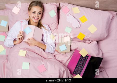 Vista dall'alto di una donna positiva che tiene la penna e il notebook vicino a libri, laptop e note adesive sul letto Foto Stock