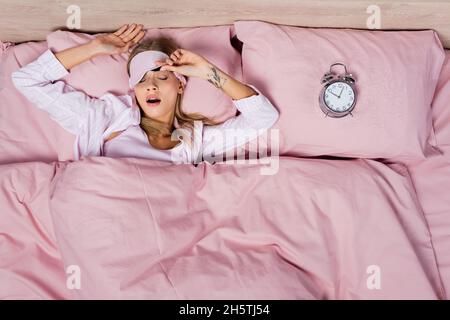 Vista dall'alto della giovane donna nel sonno maschera yawning vicino sveglia sul letto Foto Stock