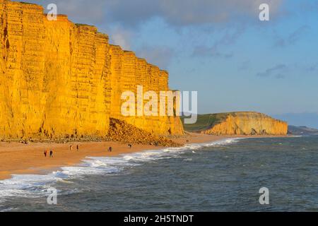 West Bay, Dorset, Regno Unito. 11 novembre 2021. Meteo Regno Unito. Camminatori sulla spiaggia godendo del sole nel tardo pomeriggio a West Bay in Dorset come il cielo chiaro. La spiaggia è attualmente bloccata da una cascata di rocce dopo che una sezione della scogliera è crollata martedì 9 novembre. Picture Credit: Graham Hunt/Alamy Live News Foto Stock