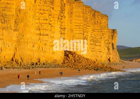 West Bay, Dorset, Regno Unito. 11 novembre 2021. Meteo Regno Unito. Camminatori sulla spiaggia godendo del sole nel tardo pomeriggio a West Bay in Dorset come il cielo chiaro. La spiaggia è attualmente bloccata da una cascata di rocce dopo che una sezione della scogliera è crollata martedì 9 novembre. Picture Credit: Graham Hunt/Alamy Live News Foto Stock