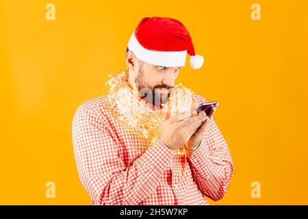 Su uno sfondo giallo, un uomo in una camicia rossa a quadri e il cappello di Babbo Natale tiene uno smartphone davanti a lui con due mani Foto Stock