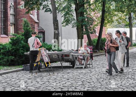 Uomo maturo, soffiatore di vetro, seduto al tavolo nel parco, in processo di lavorazione del vetro artistico. Turisti che passano vicino sono interessanti in miniature di vetro Foto Stock