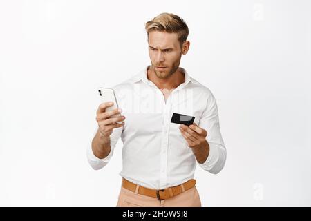 Uomo adulto confuso che guarda lo schermo del telefono cellulare, tenendo la carta di credito, in piedi su sfondo bianco Foto Stock