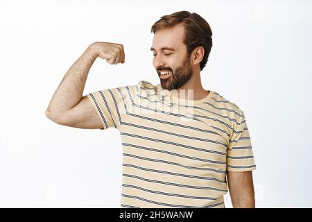 Ritratto di uomo sorridente compiaciuto guardando i muscoli del braccio, flettendo bicipiti con espressione soddisfatto del volto. Concetto di allenamento e di palestra Foto Stock