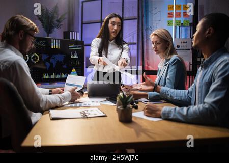 Finanziatori multietnici che discutono di una soluzione strategica per lo sviluppo aziendale mentre si siedono in ufficio. Gruppo di quattro colleghi che lavorano con carte e gadget moderni. Foto Stock