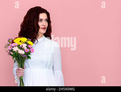 Donna che tiene bouquet di fiori guardando verso sinistra su sfondo rosa. Ritratto di bella donna che tiene bouquet di fiori in una mano. Foto Stock