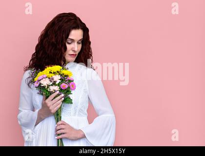 Donna che tiene i fiori al torace guardando giù su sfondo rosa. Ritratto di donna pensiva triste in abito bianco che tiene bouquet al suo petto. Foto Stock