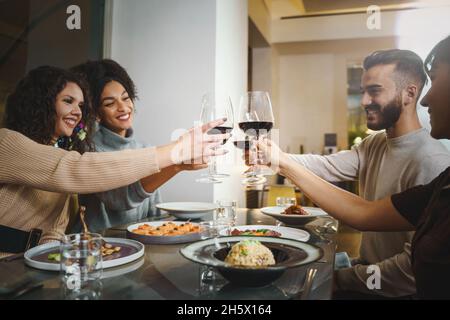 Gruppo di amici multirazziali che accecano i wineglasses - riunione degli amici che tostano il vino rosso al ristorante Foto Stock