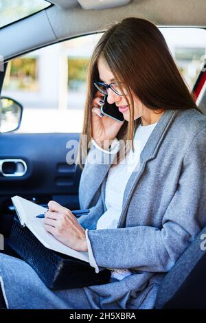 Imprenditore femminile occupato seduto sul sedile del passeggero in auto e prendere appunti in blocco durante la telefonata di lavoro su smartphone Foto Stock