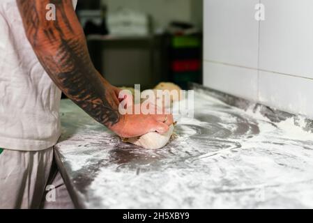 Vista laterale del panettiere maschio tatuato di raccolto in polo bianco che impastano l'impasto con le mani mentre si trova al banco di metallo in cucina della moderna panetteria Foto Stock