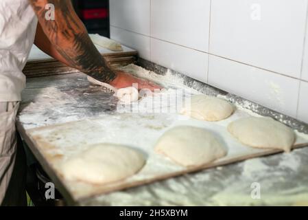 Vista laterale del panettiere maschio tatuato di raccolto in polo bianco che impastano l'impasto con le mani mentre si trova al banco di metallo in cucina della moderna panetteria Foto Stock