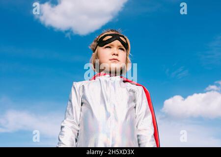 Da sotto cute capretto che indossa il costume del supereroe della mascheratura e la maschera dell'occhio che si levano in piedi contro il cielo blu e che guardano via Foto Stock