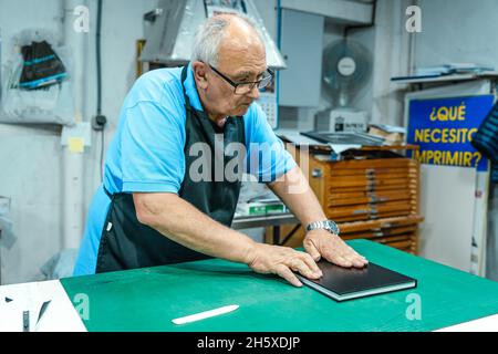 Artigiano maschio di mezza età in grembiule che fa rilegatura per libro in piedi a tavola in laboratorio di stampa Foto Stock