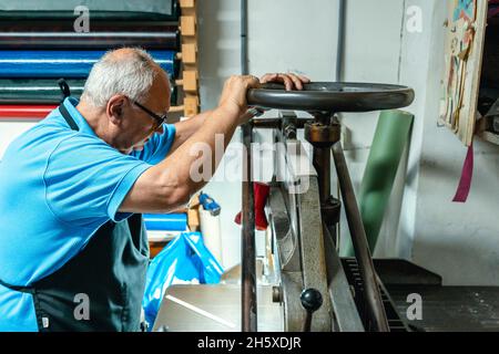 Vista laterale di un artigiano maschile anziano focalizzato in abiti casual e ruota di filatura grembiule mentre si utilizza la macchina per la legatura in officina di stampa Foto Stock