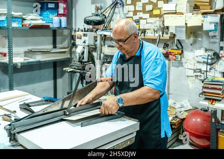 Artigiano maschile anziano focalizzato in grembiule e occhiali tagliando pezzo di cartone con macchina mentre si lavora in tipografia Foto Stock