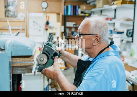 Vista laterale del carpentiere maschile concentrato con capelli bianchi in grembiule e occhiali tagliando tavole di legno con sega circolare durante il lavoro in Falegnameria Foto Stock