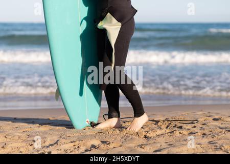 Vista laterale di una surfista femminile inriconoscibile e tagliata in muta con tavola da surf in piedi che guarda via sulla riva bagnata dal mare ondeggiante Foto Stock