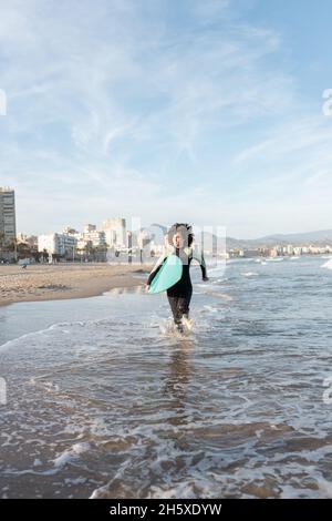 Giovane surfista donna premurosa in muta con tavola da surf che corre guardando via sulla riva bagnata dal mare ondeggiante Foto Stock