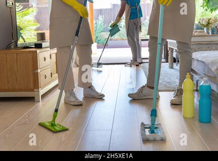 I colleghi sono tagliati e irriconoscibili in grembiuli e guanti che puliscono il pavimento in parquet con mocio e scope durante il lavoro in un soggiorno moderno e spazioso Foto Stock