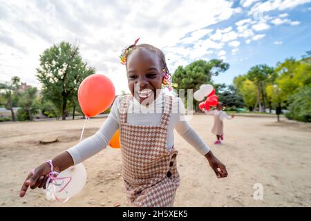 Sorelline afroamericane felici in abiti simili in piedi con palloncini colorati in mani su erba verde nel parco alla luce del giorno Foto Stock