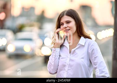 Conversazione telefonica vicino autostrada con traffico intenso, la giovane donna utilizza smartphone sulla strada della città la sera d'estate. Foto Stock