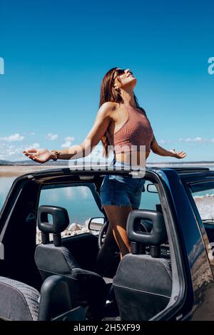 Vista laterale del giovane viaggiatore tatuato femmina in cima e pantaloncini in denim in piedi in SUV cabriolet auto parcheggiato su spiaggia sabbiosa e ammirare il pittoresco mare Foto Stock