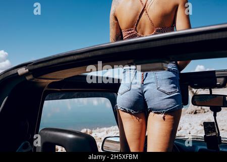 Vista posteriore di una giovane e irriconoscibile viaggiatore tatuato in cima e pantaloncini in denim in piedi in SUV cabriolet auto parcheggiata sulla spiaggia sabbiosa e ammirare Foto Stock