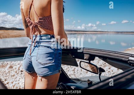 Vista posteriore di una giovane e irriconoscibile viaggiatore tatuato in cima e pantaloncini in denim in piedi in SUV cabriolet auto parcheggiata sulla spiaggia sabbiosa e ammirare Foto Stock