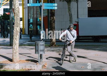 Intera lunghezza di lavoratore maschio bearded in casco che attraversa strada asfaltata con la bicicletta sulla strada di lavoro Foto Stock