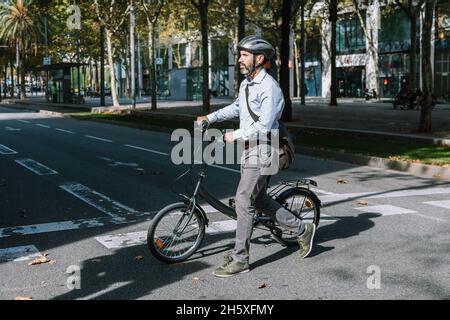 Intera lunghezza di lavoratore maschio bearded in casco che attraversa strada asfaltata con la bicicletta sulla strada di lavoro Foto Stock